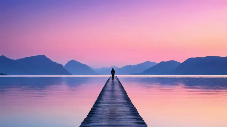 A serene desktop image of a solitary figure standing on a causeway at twilight, reflected in calm water, with a deep blue and pink horizon.